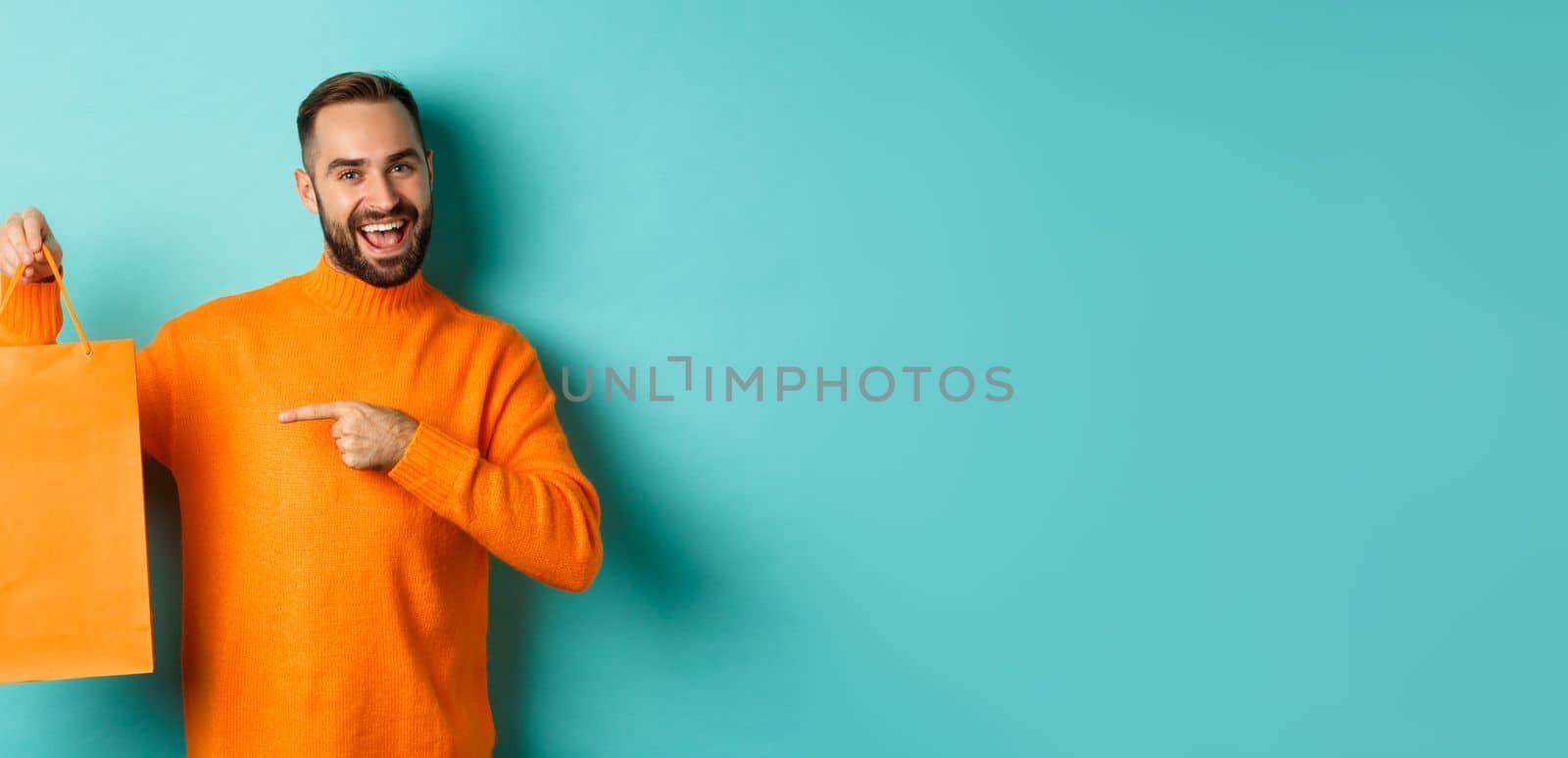 Satisfied male customer pointing at orange shopping bag, recommending store, smiling pleased, standing over turquoise background.