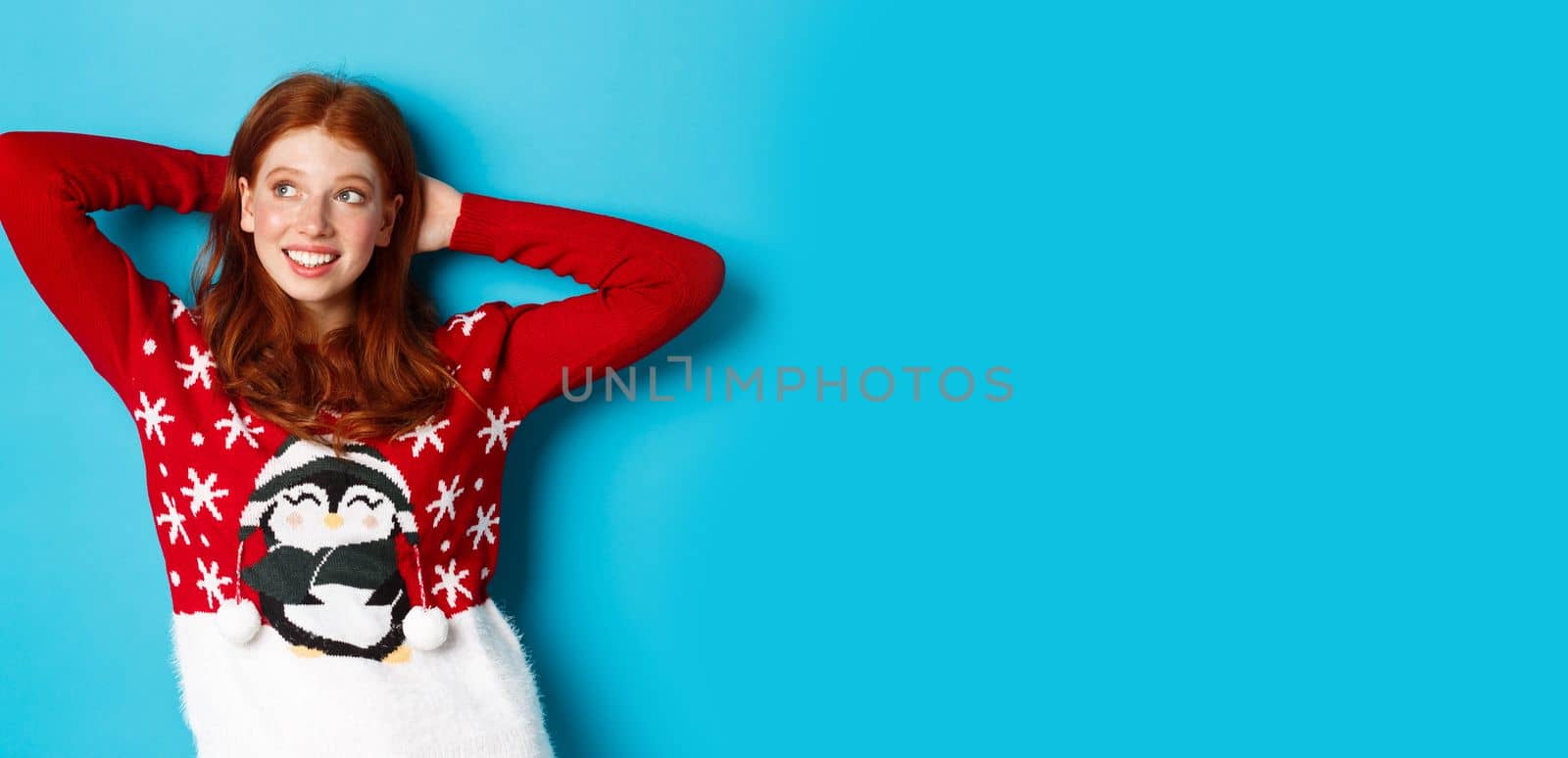 Winter holidays and celebration concept. Relaxed teenage girl holding hands behind head and looking left at promo, resting on Christmas Eve, standing over blue background by Benzoix