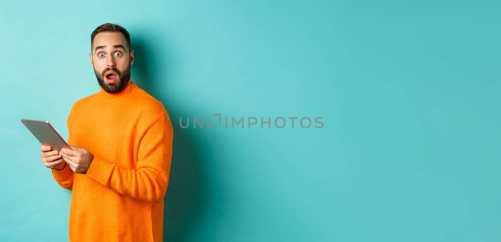 Image of male model in orange sweater using digital tablet, looking surprised, standing over light blue background.