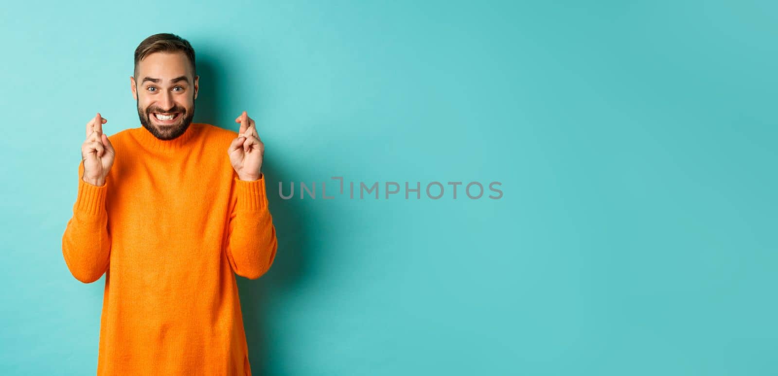 Excited man with beard, making a wish, holding fingers crossed for good luck and smiling, standing over light blue background.