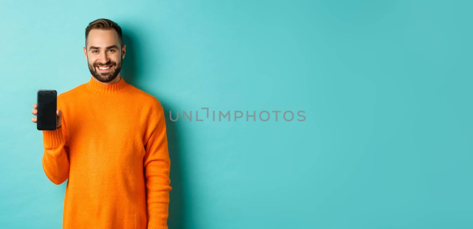 Handsome bearded guy in orange sweater, showing smartphone screen and smiling, showing promo online, turquoise background by Benzoix