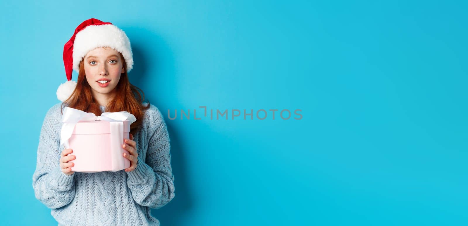 Winter holidays and Christmas Eve concept. Cute redhead girl wearing Santa hat, holding New Year gift and looking at camera, standing against blue background.