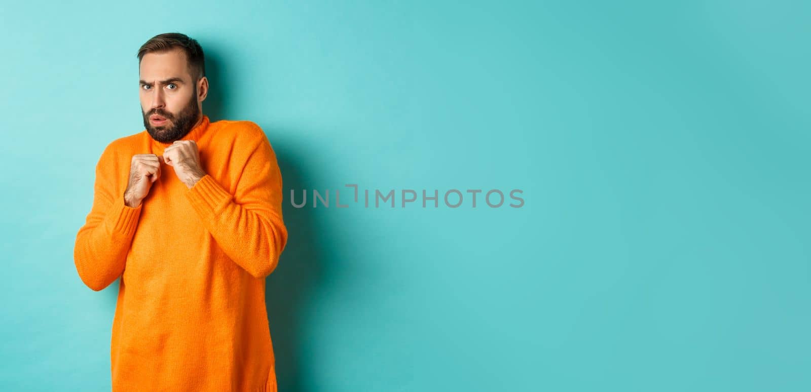 Scared guy jumping startled, looking at something scary, standing in orange sweater, studio background.