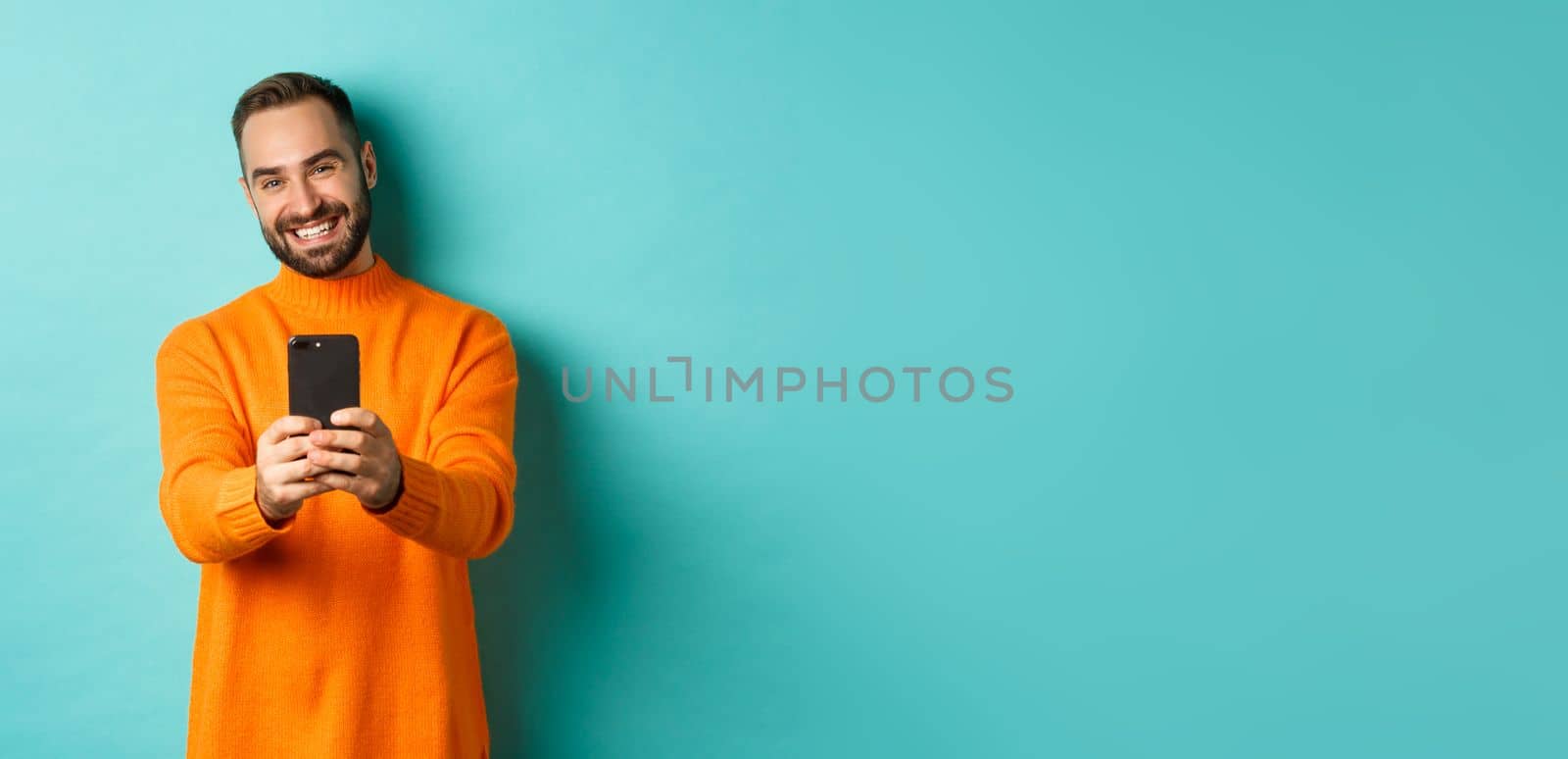 Happy handsome man taking photo on mobile phone, take pictures with smartphone camera, standing over light blue background.