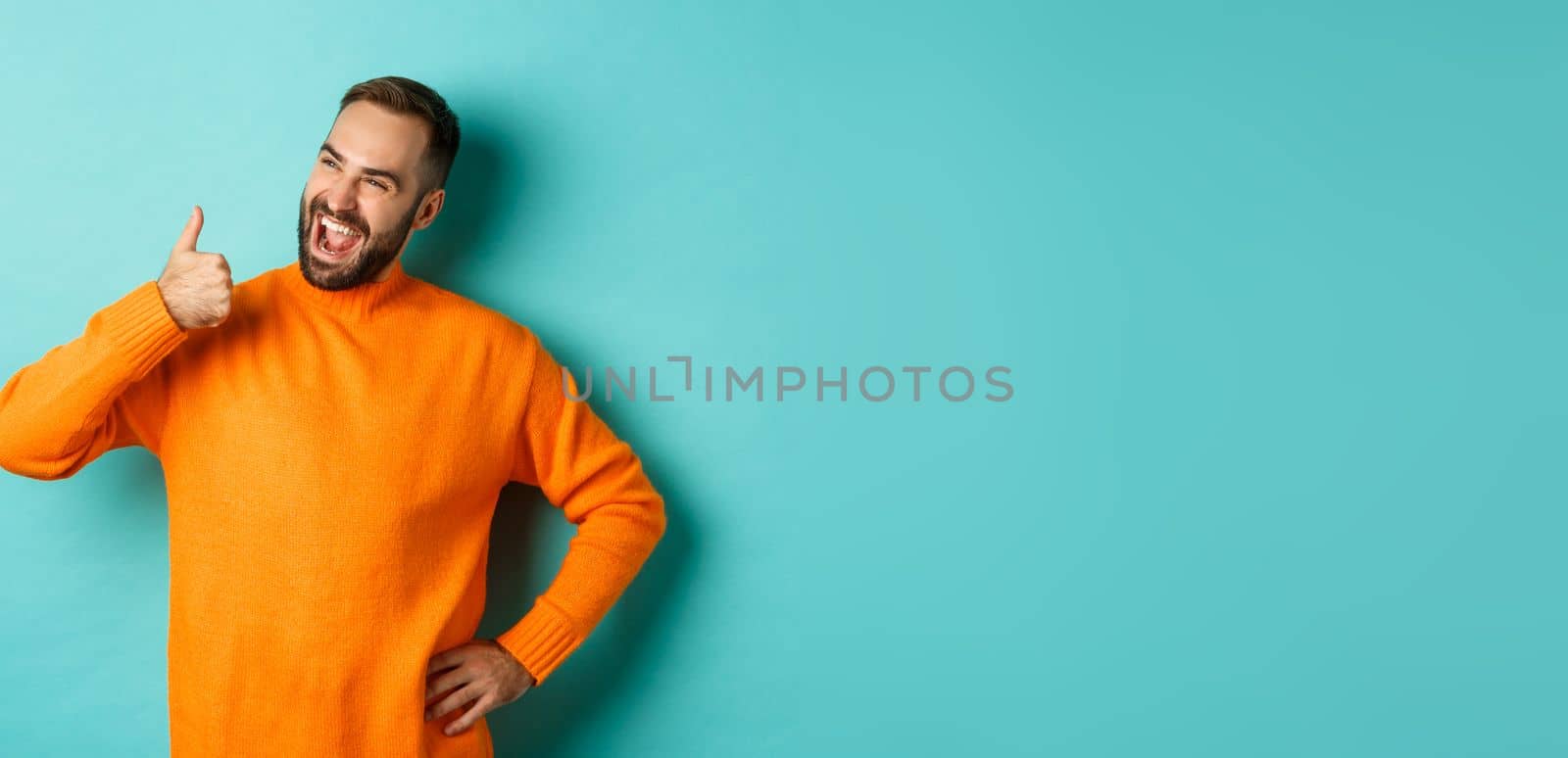Happy satisfied male model showing thumb up, looking pleased at upper left corner logo, approve and recommend product, standing over light blue background.