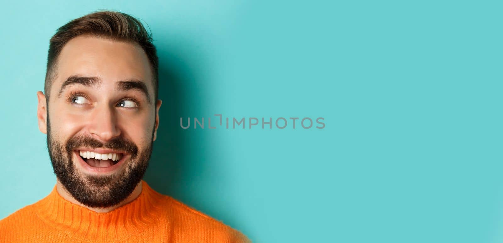 Headshot of handsome caucasian man looking at upper left corner logo and smiling pleased, checking out promo offer, standing over turquoise background by Benzoix
