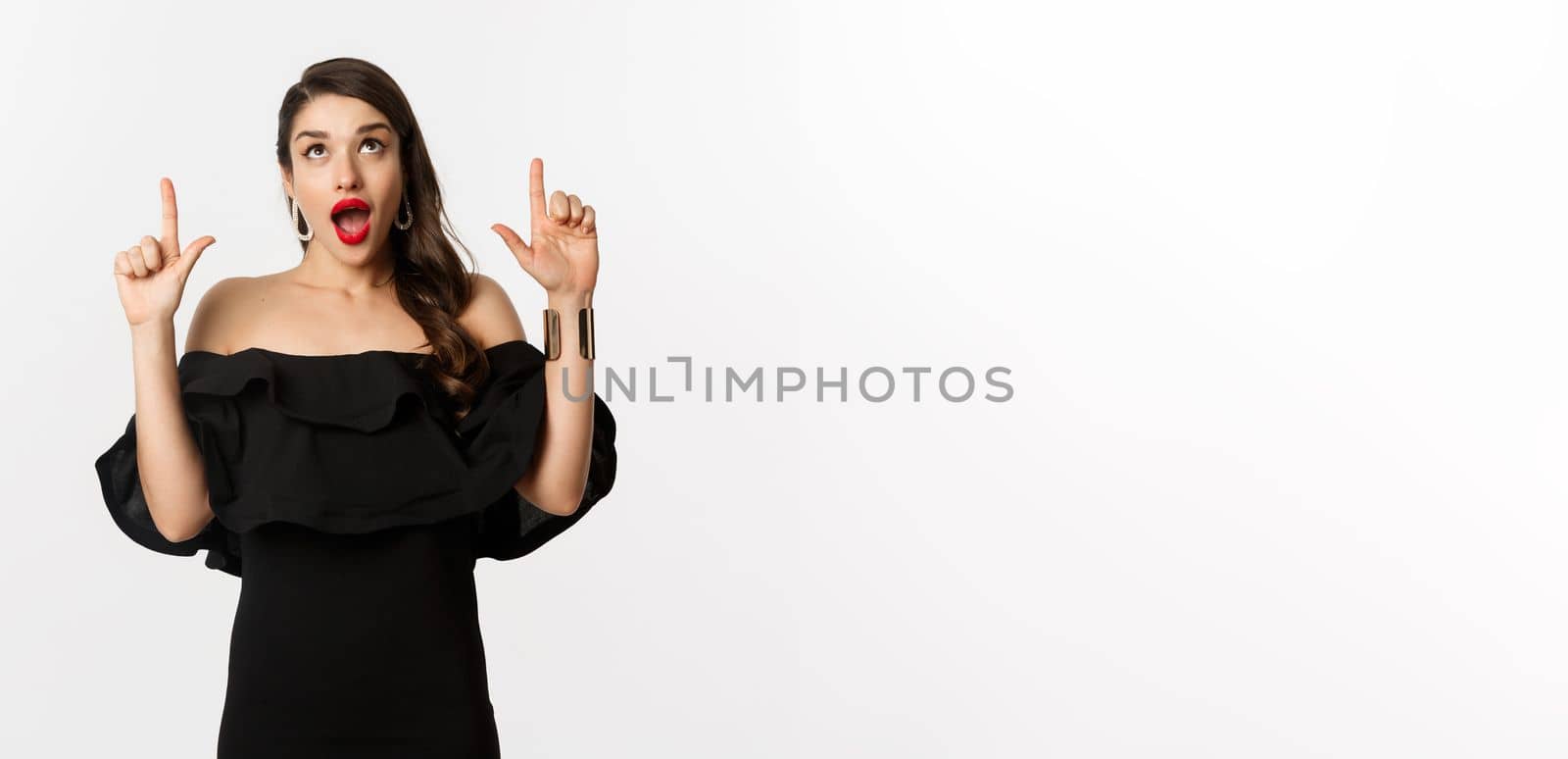 Fashion and beauty. Amused woman in black dress looking and pointing fingers up at promo offer, standing over white background.