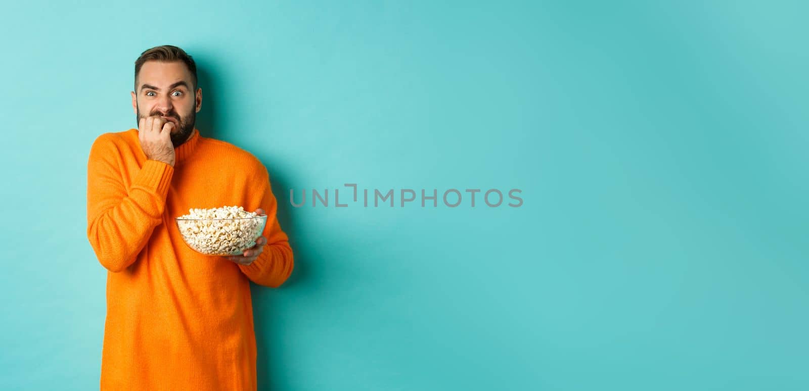 Image of scared young man watching horror movie, biting fist and looking terrified, holding bowl of popcorn, standing over turquoise background by Benzoix