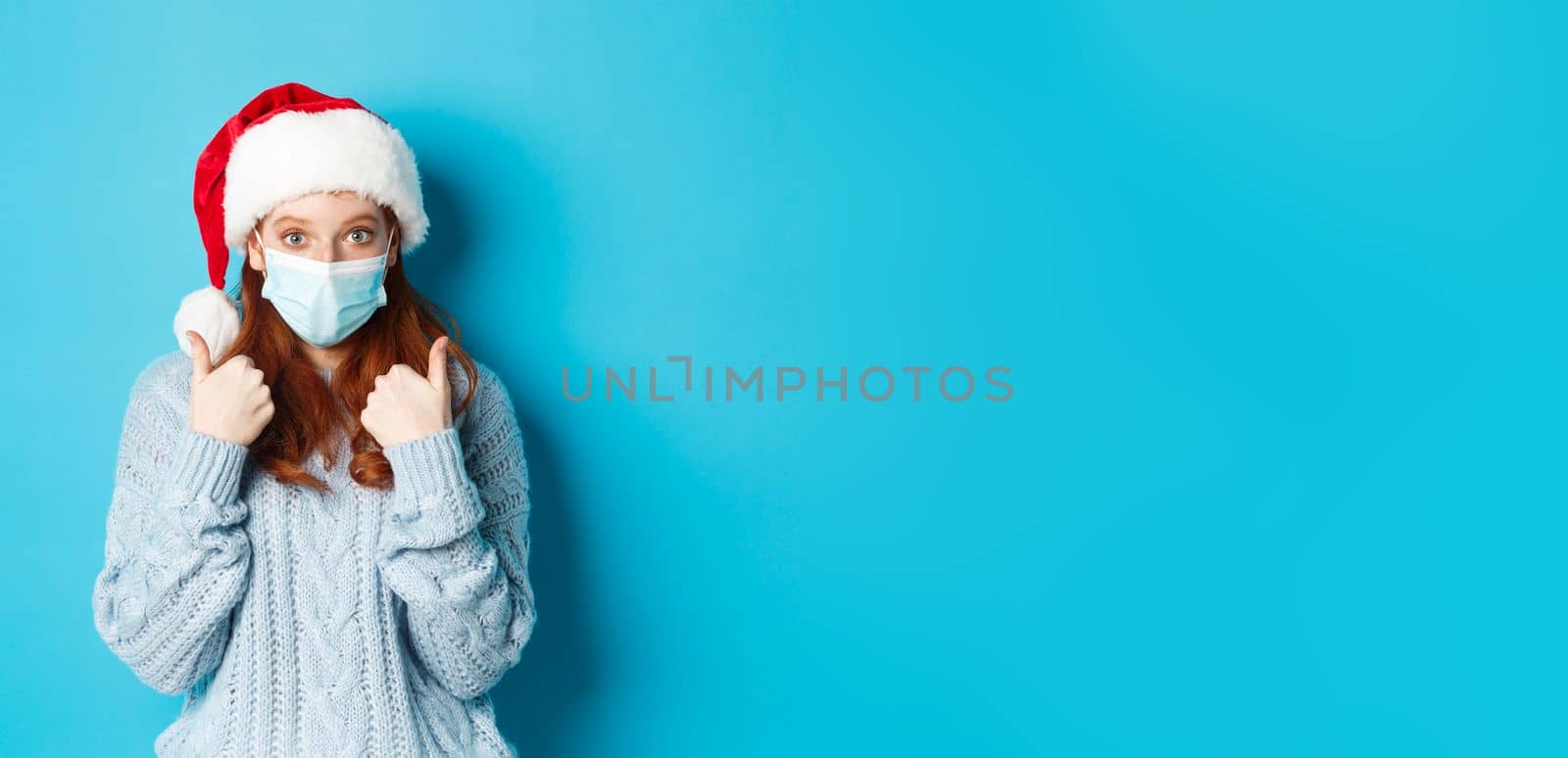 Christmas, quarantine and covid-19 concept. Cute teen redhead girl in santa hat and sweater, wearing face mask from coronavirus, showing thumbs up, standing over blue background by Benzoix