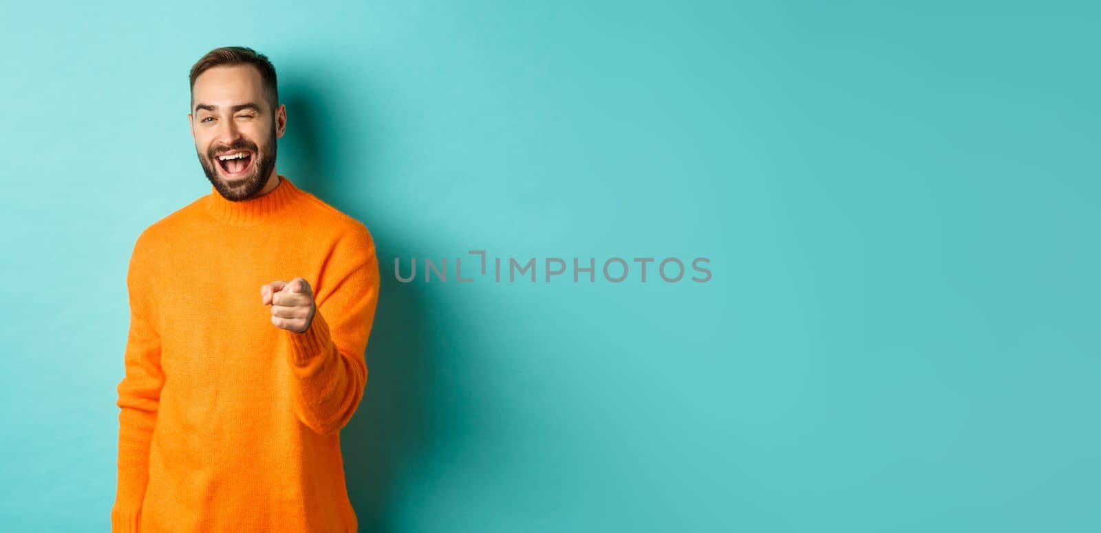 Cheeky attractive man congrats you, praising good job, winking and pointing at camera, standing over light blue background.