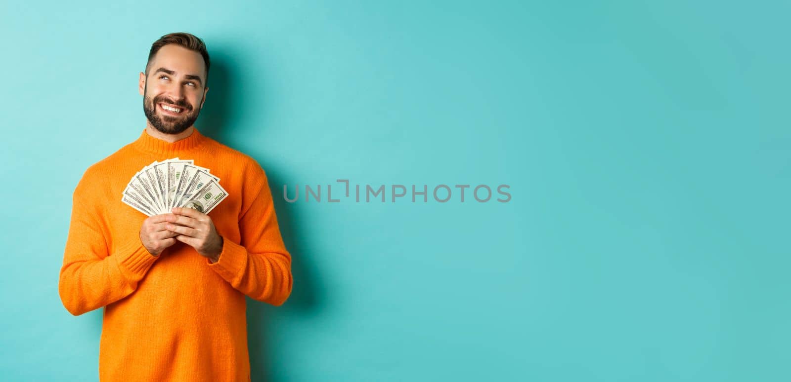 Man thinking about shopping, holding money and smiling satisfied, looking at upper left corner, standing over light blue background.