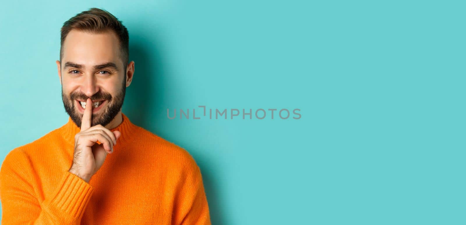 Close-up of handsome smiling man hushing, telling secret, standing over light blue background by Benzoix