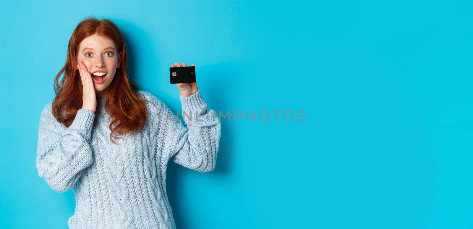 Cute redhead girl in sweater showing credit card, smiling at camera, standing over blue background by Benzoix