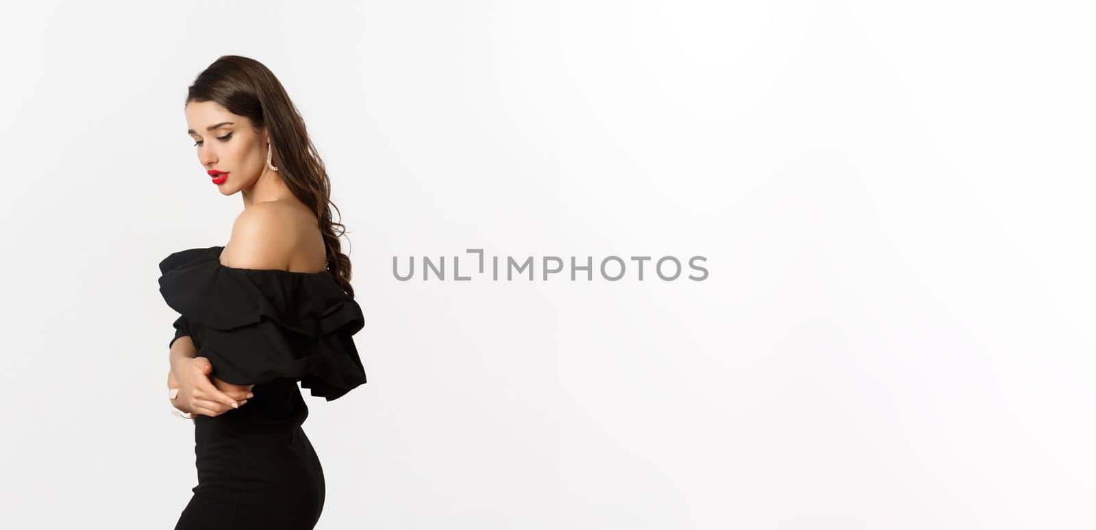 Profile of sensual brunette woman in black elegant dress, looking down, standing over white background.