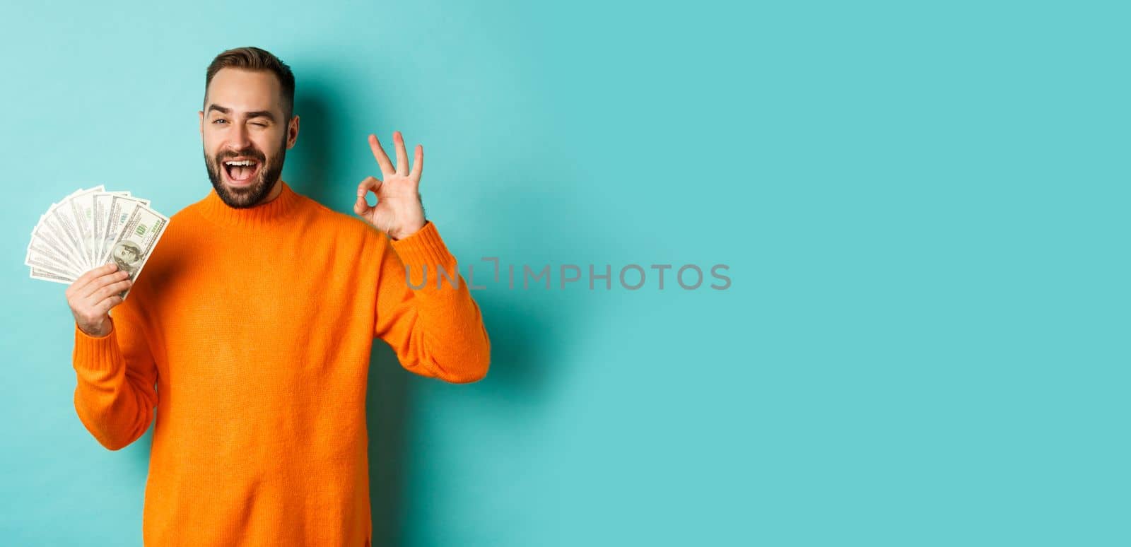 Shopping. Excited guy holding money, showing ok sign and winking, standing over light blue background. Copy space