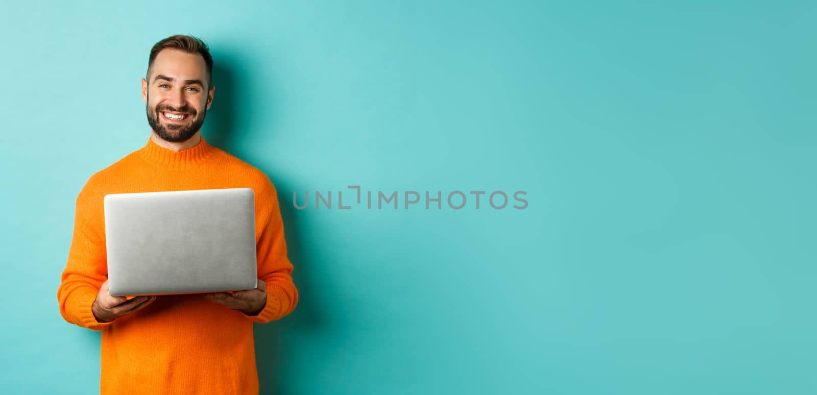 Handsome freelancer working on laptop and smiling, standing in orange sweater over light blue background by Benzoix