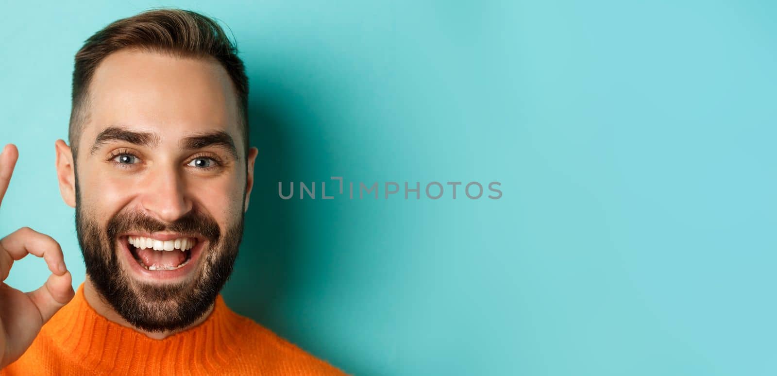 Close-up of confident and happy man with beard showing okay sign, approve and like, giving positive reply or agree, standing over turquoise background.