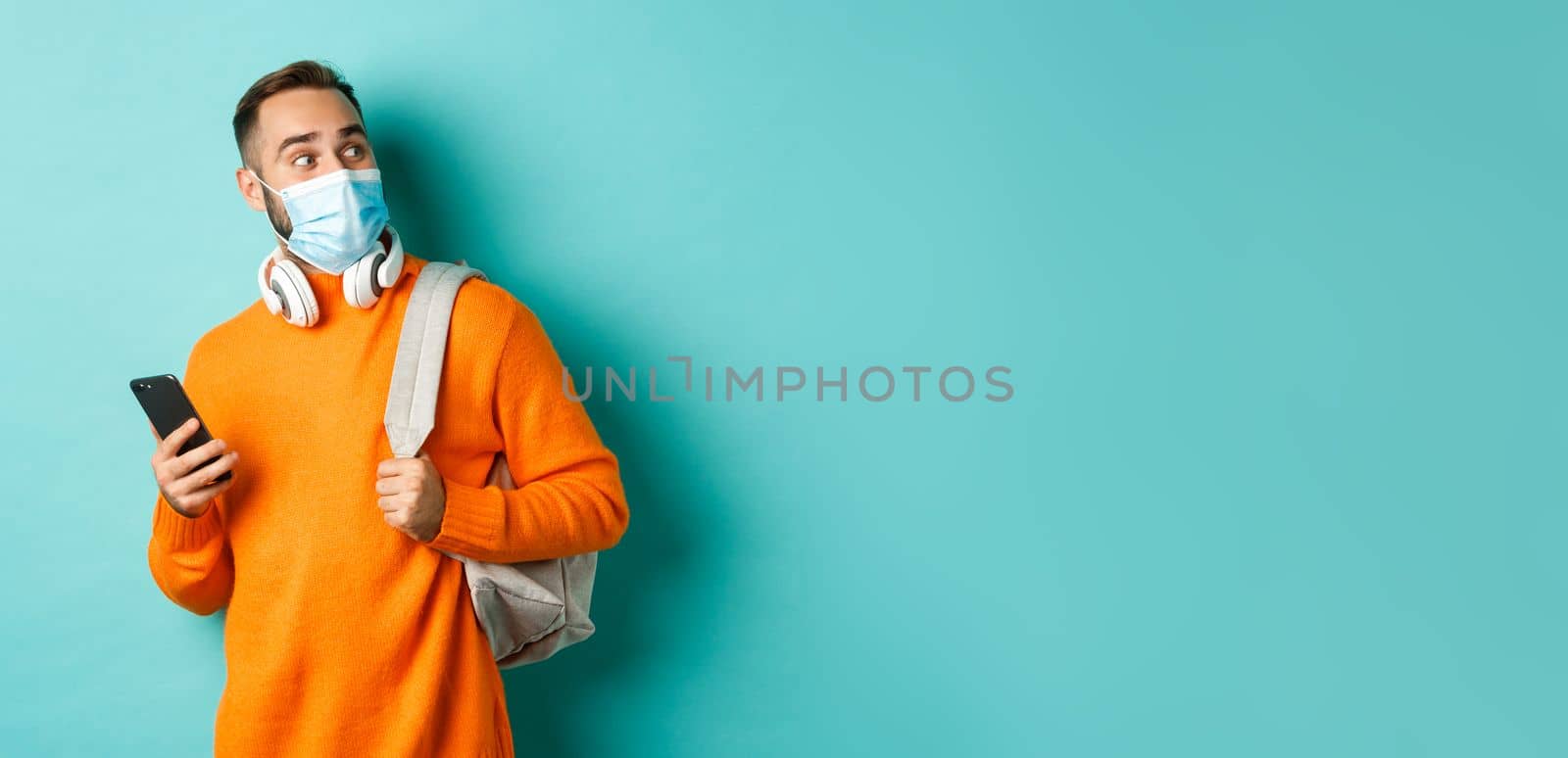 Young man in face mask using mobile phone, holding backpack, staring right amazed, standing against light blue background.