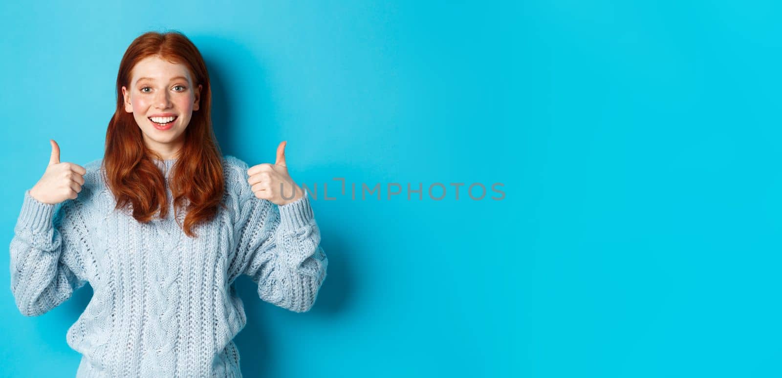 Cheerful teen girl with red hair, showing thumbs up in approval, like and praise gesture, standing over blue background.