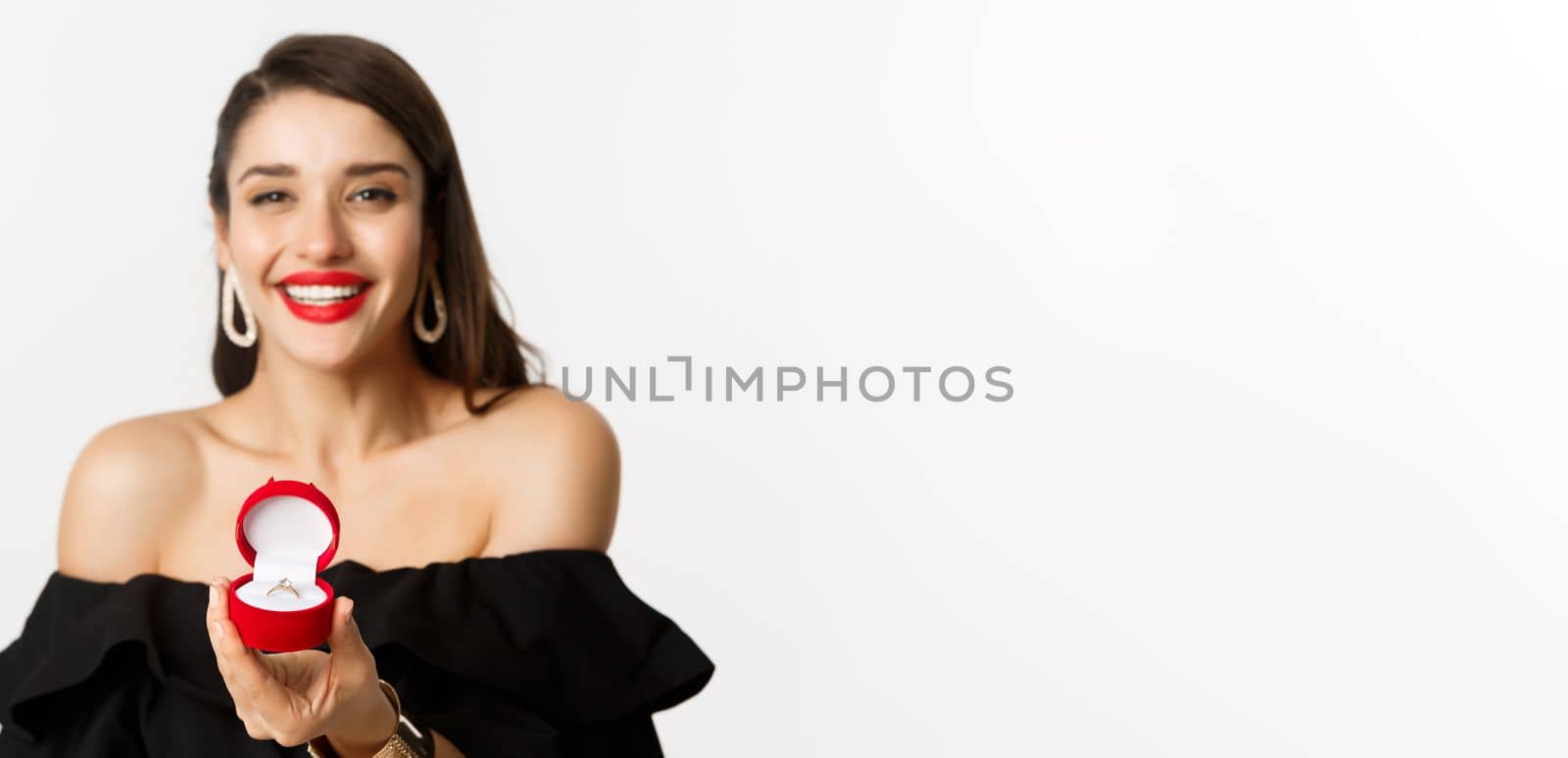 Fashion and beauty concept. Close-up of gorgeous brunette woman with red lips, black dress, showing engagement ring in box and smiling, getting married, standing over white background.