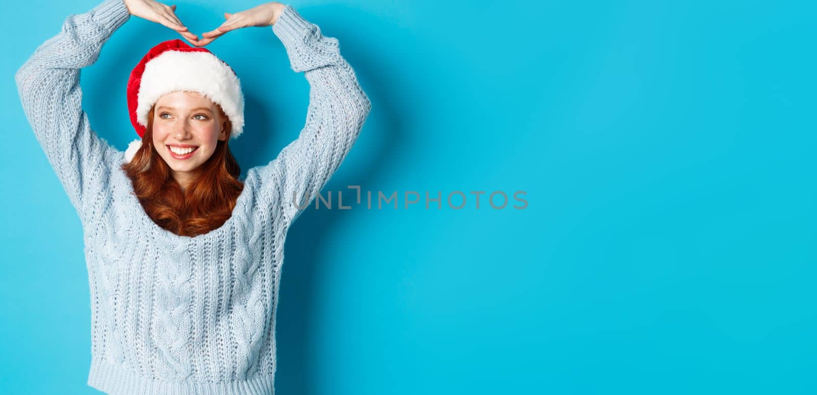 Winter holidays and Christmas Eve concept. Cute redhead teen girl in santa hat and sweater, making heart sign and smiling, wishing merry xmas, standing over blue background.