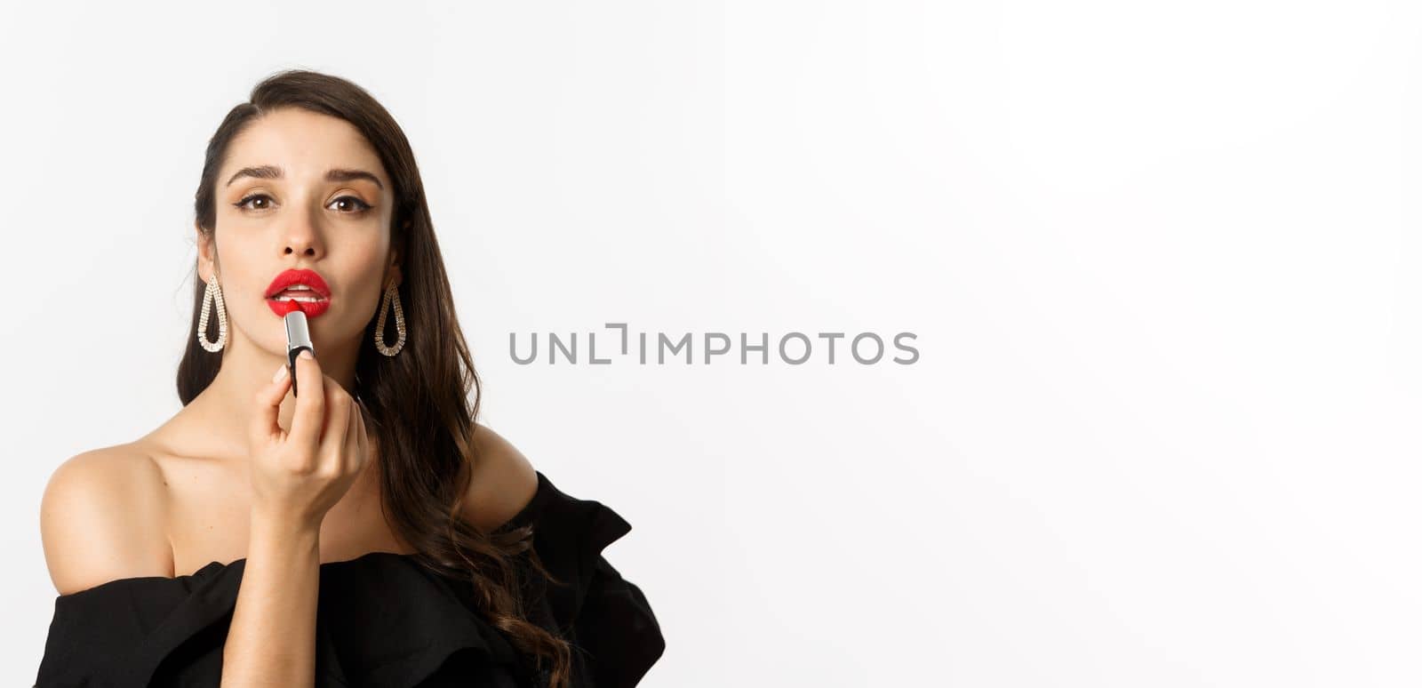 Close-up of beautiful woman applying red lipstick on lips, looking at camera like mirror, standing in black dress over white background by Benzoix