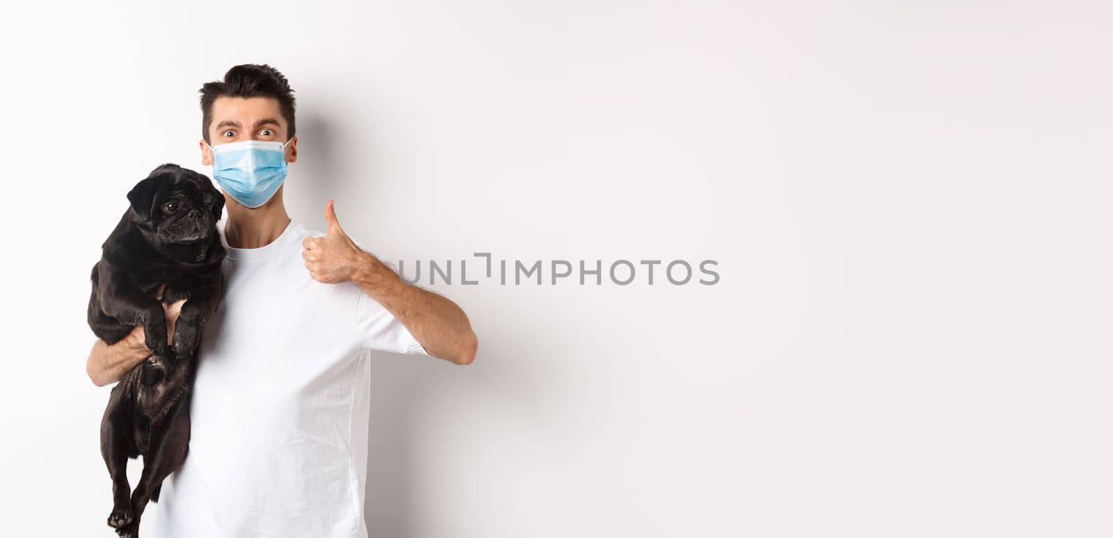 Covid-19, animals and quarantine concept. Young man in medical mask holding cute black pug dog, showing thumb up, like and approve, standing over white background by Benzoix