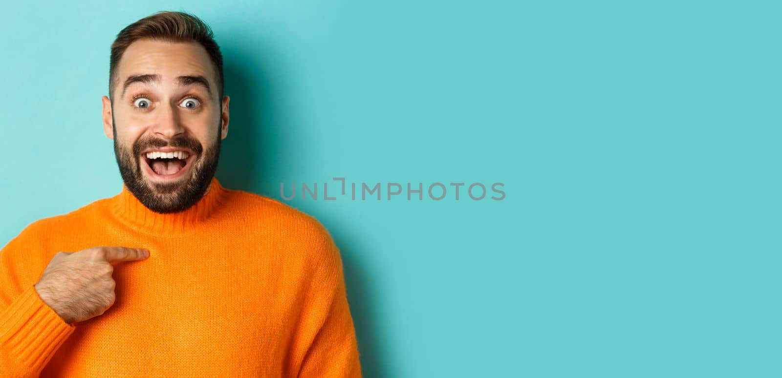 Close-up of happy young man pointing at himself with surprise and excitement, standing over light blue background.