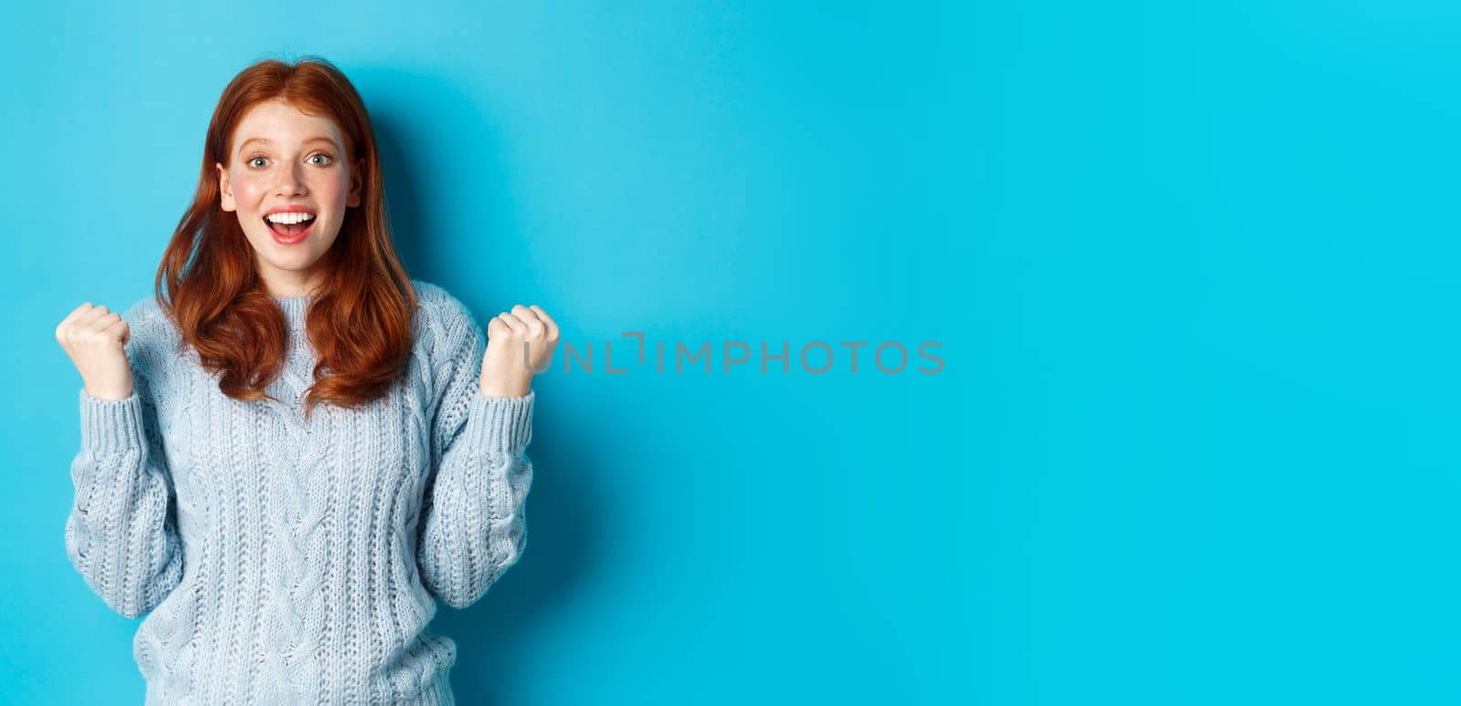 Satisfied redhead girl achieve goal and celebrating, making fist pump gesture and smiling with rejoice, triumphing of win, standing against blue background by Benzoix