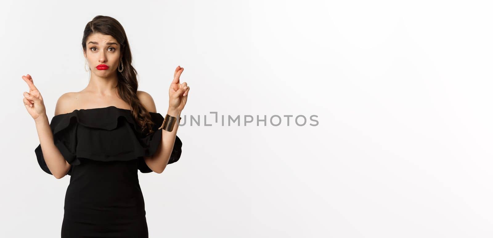 Fashion and beauty. Hopeful silly woman in black dress making wish, holding fingers crossed for good luck, standing over white background by Benzoix