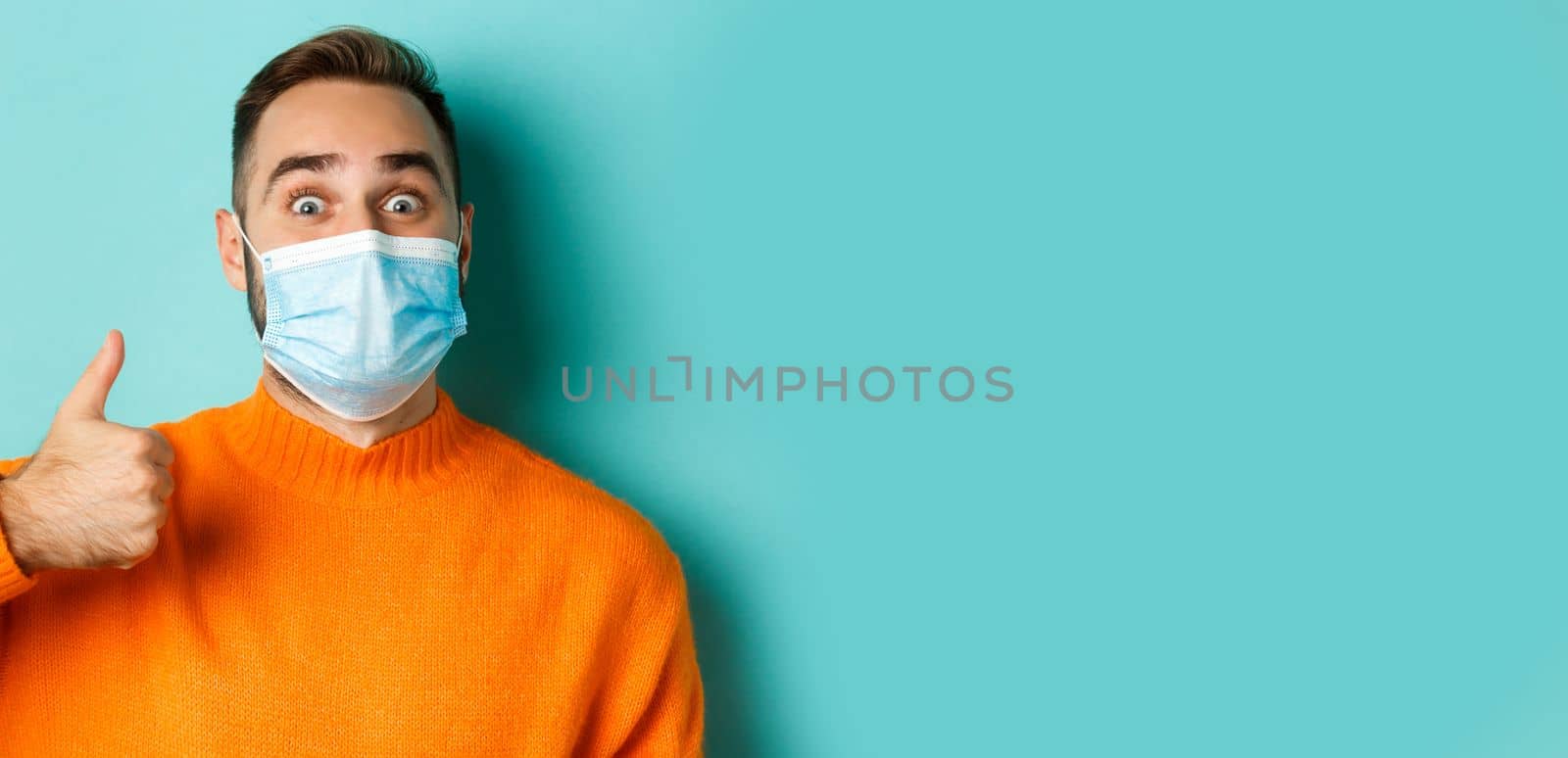 Covid-19, social distancing and quarantine concept. Close-up of cheerful man in medical mask showing thumb-up, light blue background by Benzoix