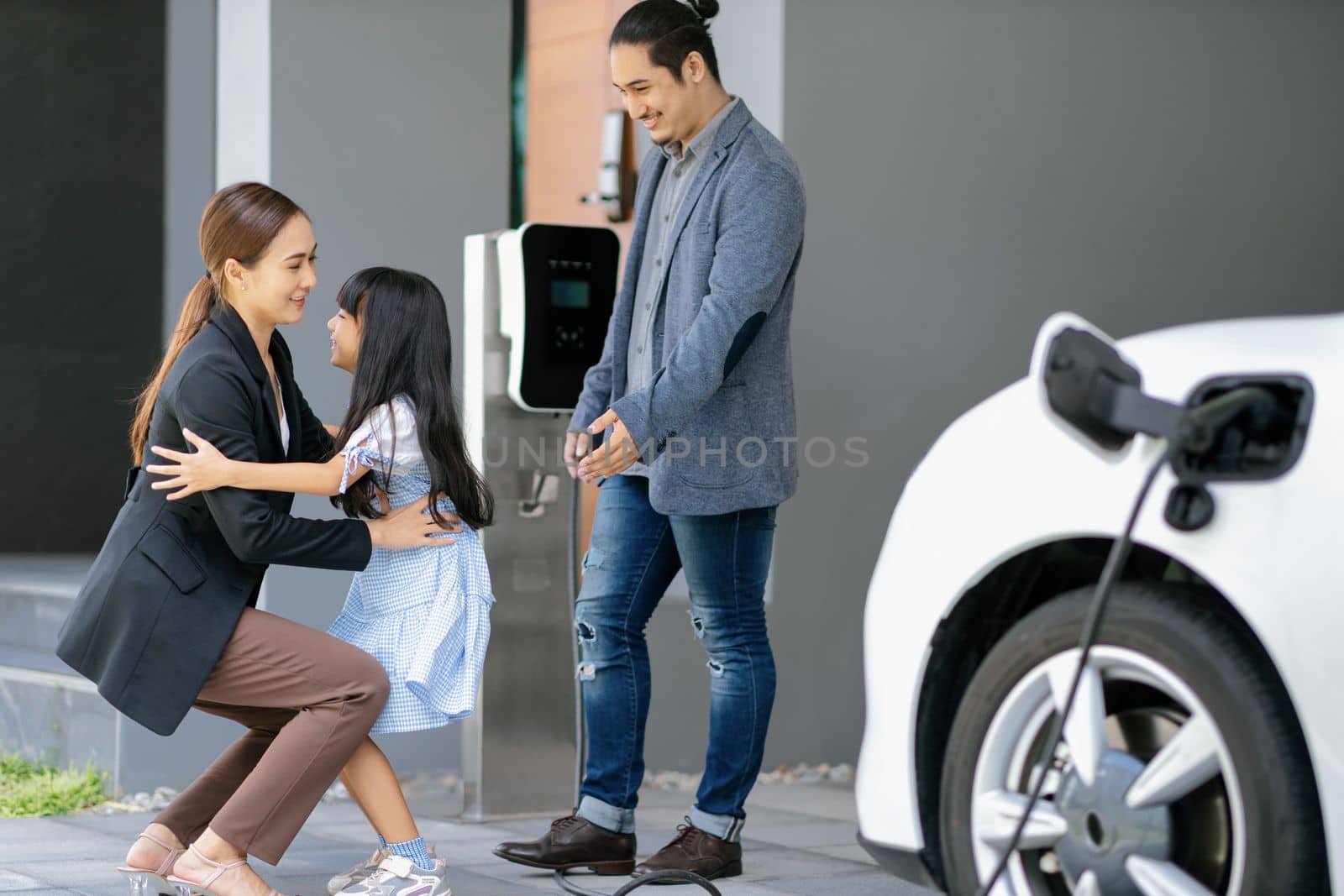 Progressive young parents and daughter with electric vehicle and home charging station. Green and clean energy from electric vehicles for healthy environment. Eco power from renewable source at home.