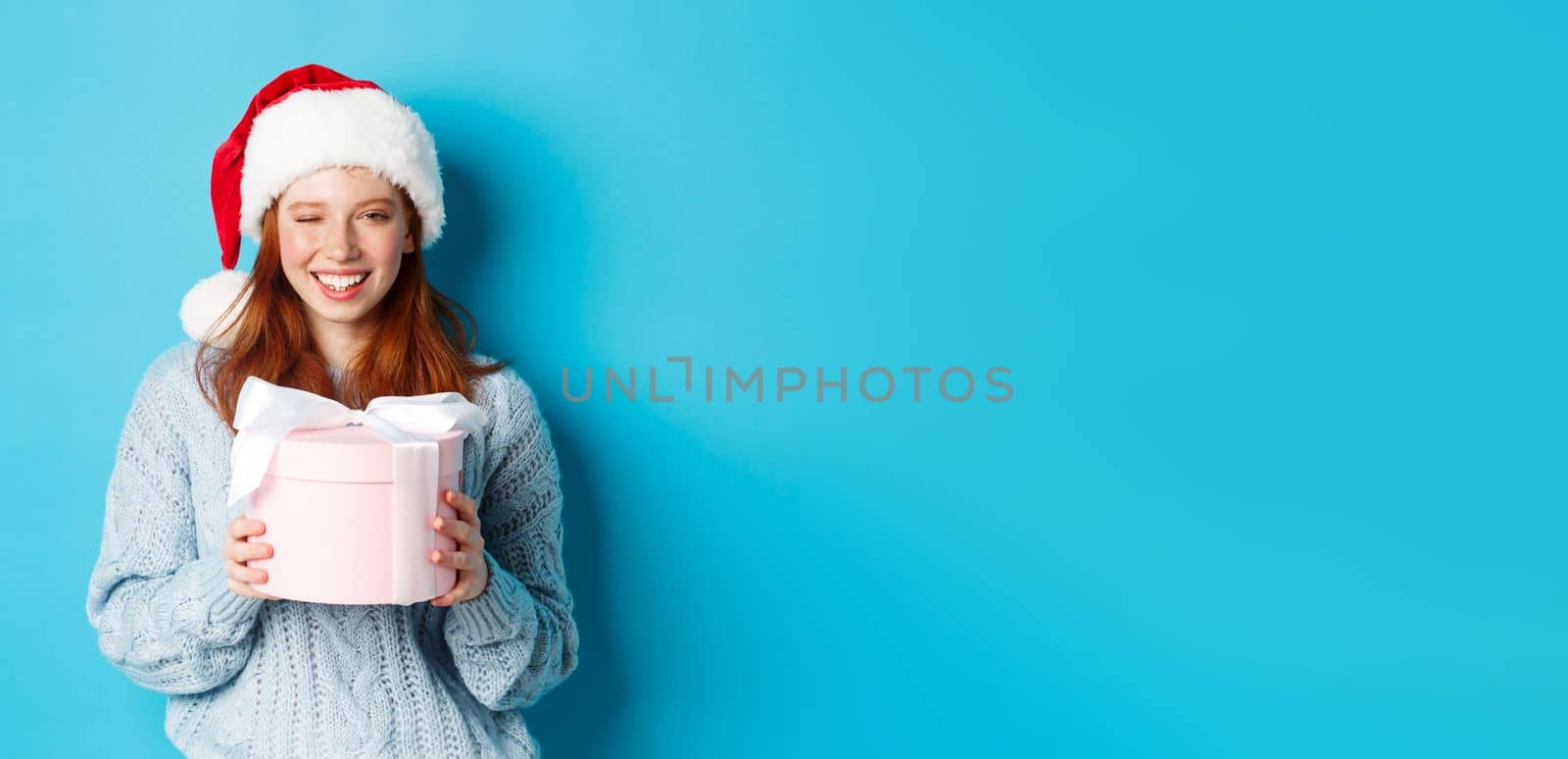 Winter holidays and Christmas Eve concept. Cute redhead girl in sweater and Santa hat, holding New Year gift and looking at camera, standing against blue background.