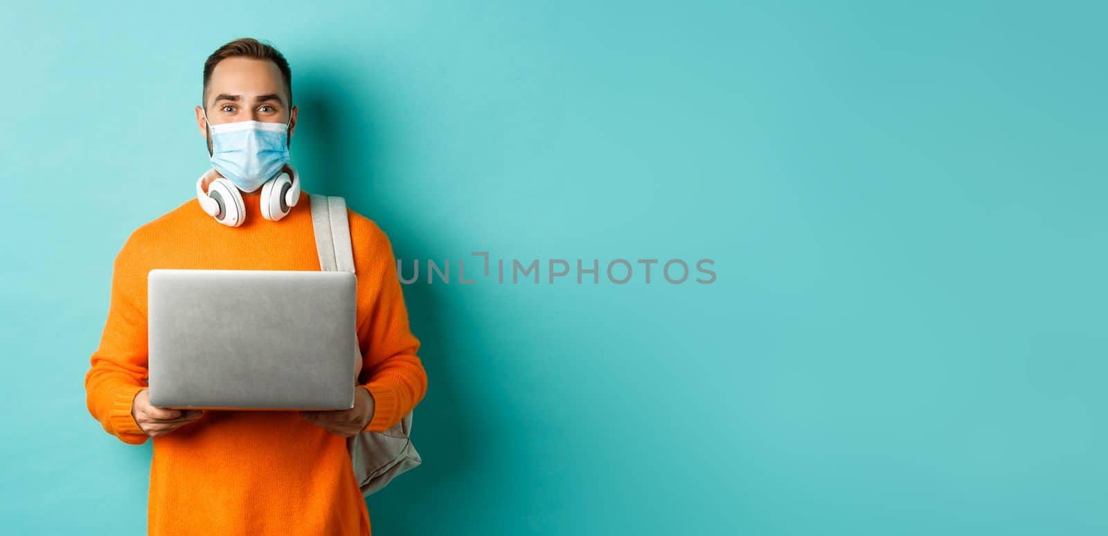 Young man in face mask using laptop and staring happy at camera, standing over light blue background.