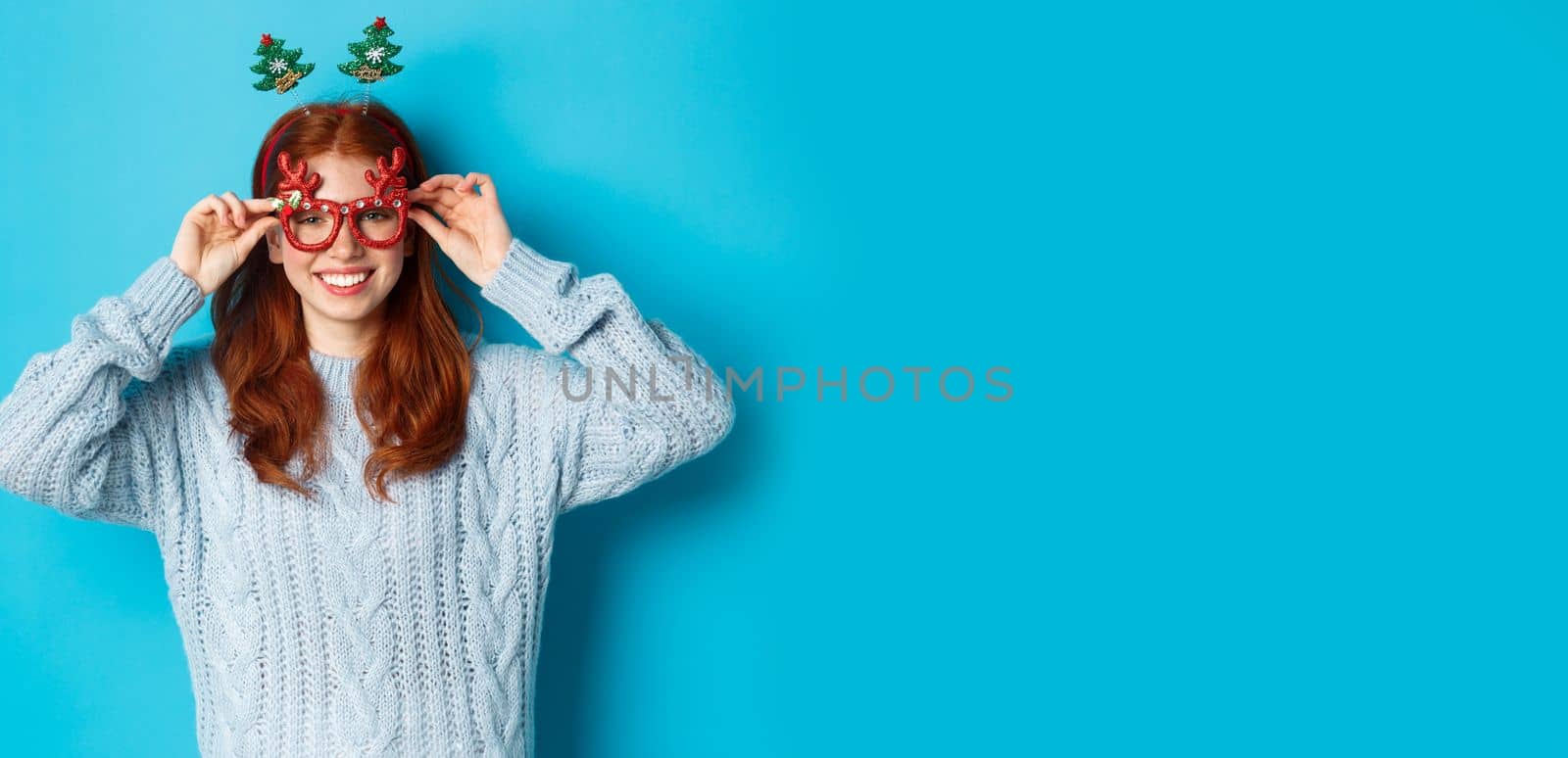 Winter holidays and Christmas sales concept. Beautiful redhead female model celebrating New Year, wearing funny party headband and glasses, smiling at camera by Benzoix