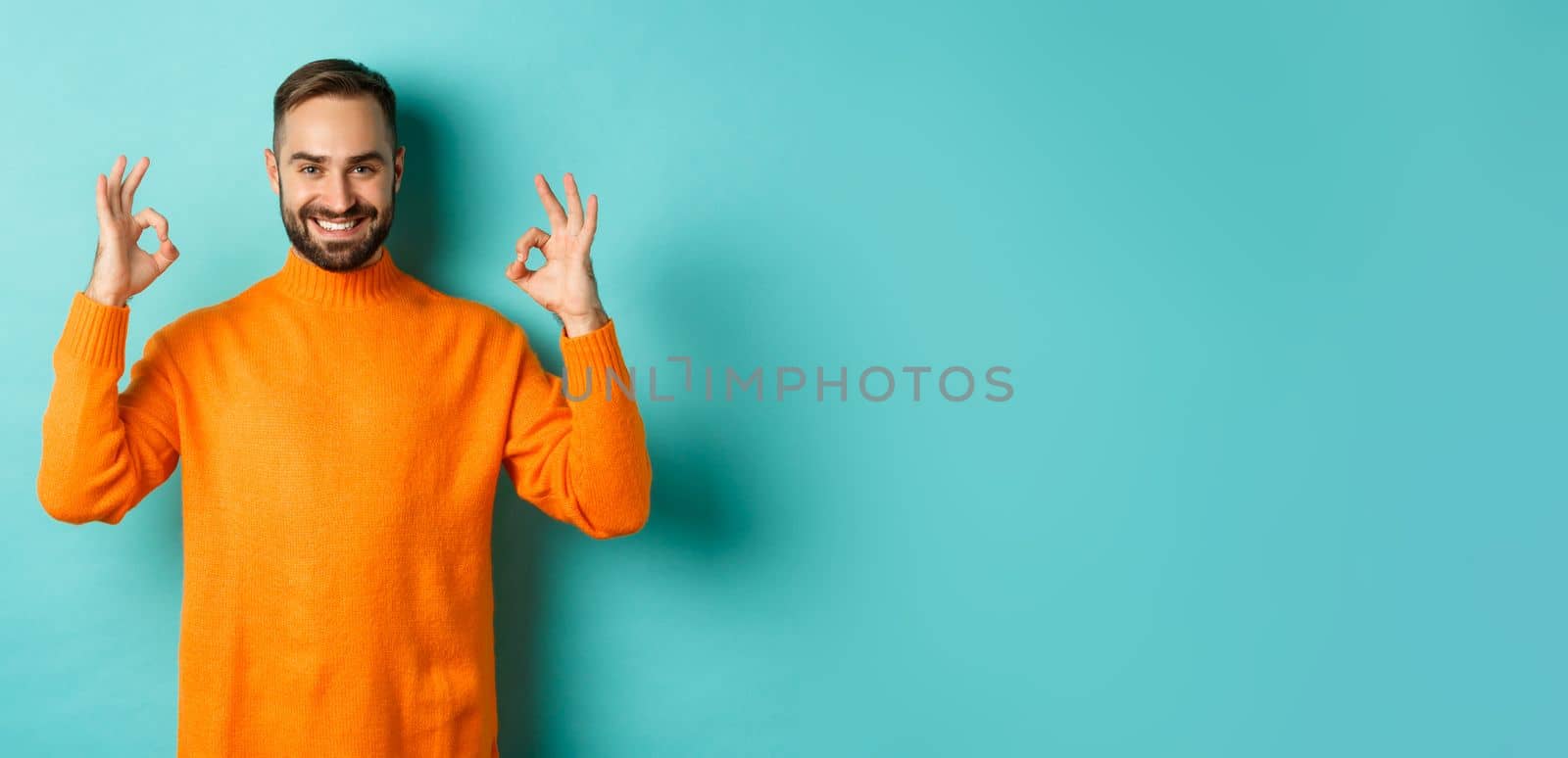 Image of confident smiling man showing okay sign, approve and agree, guarantee quality, standing over light blue background.