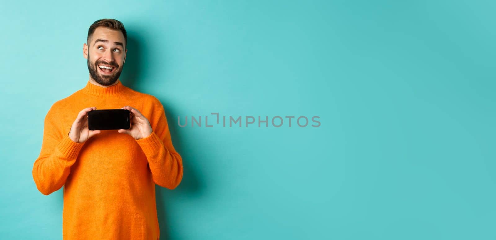 Online shopping. Young man thinking and showing smartphone screen, looking dreamy at upper left corner, standing over light blue background.