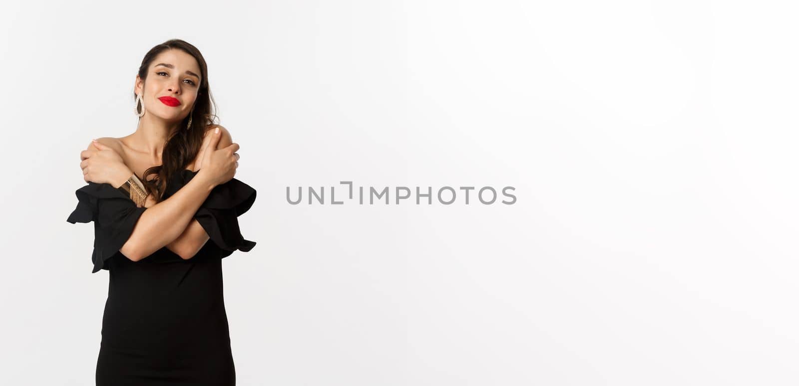 Fashion and beauty. Sensual and tender woman in black dress, embracing own body, hugging herself and smiling, standing over white background.