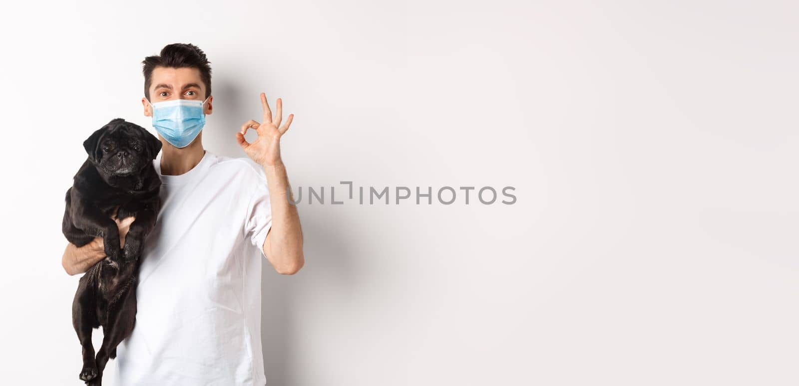 Covid-19, animals and quarantine concept. Young man in medical mask holding cute black pug dog, showing okay sign, like and approve, standing over white background.