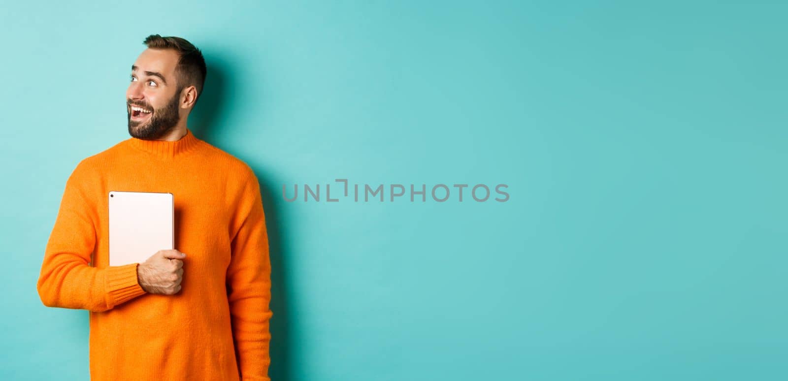Handsome man holding laptop, looking left with surprise and amazement, standing in orange sweater against light blue background.