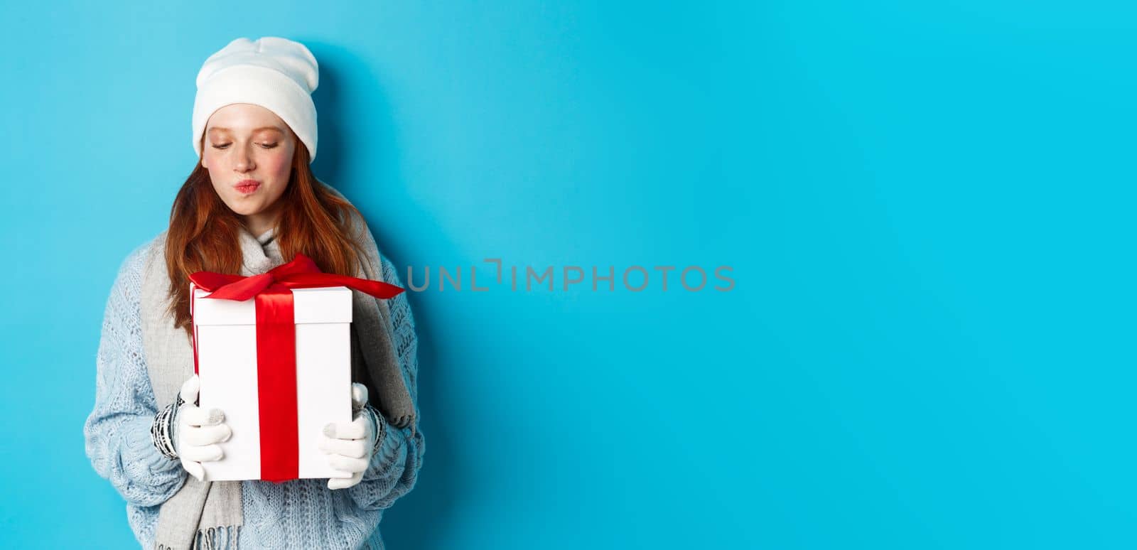 Winter holidays and Christmas sales concept. Intrigued redhead girl holding present, curiously staring at box with gift, trying guess what inside, standing over blue background.