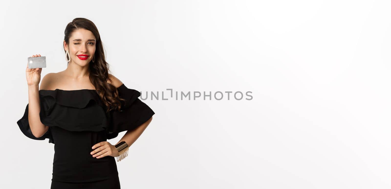 Beauty and shopping concept. Fashionable woman in black dress, wearing makeup, winking at camera and showing credit card, standing over white background.