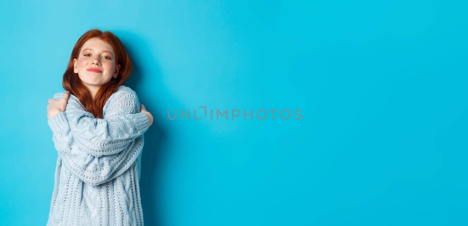 Happy cute redhead girl hugging herself, wearing comfortable and warm sweater, smiling at camera, standing over blue background by Benzoix