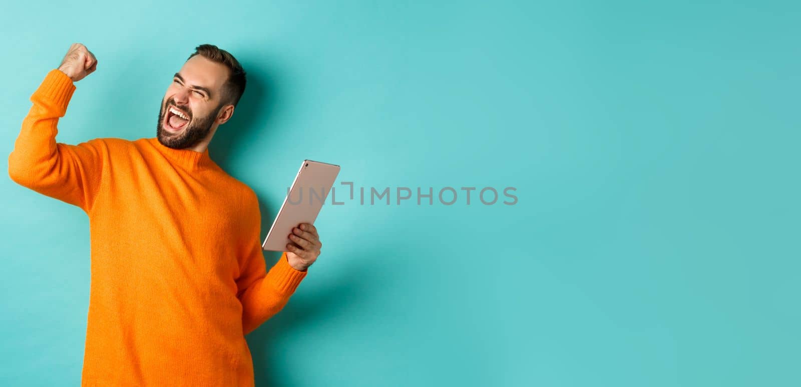 Cheerful winning man holding digital tablet, rejoicing and celebrating victory in game, making fist pump gesture, standing over light blue background by Benzoix