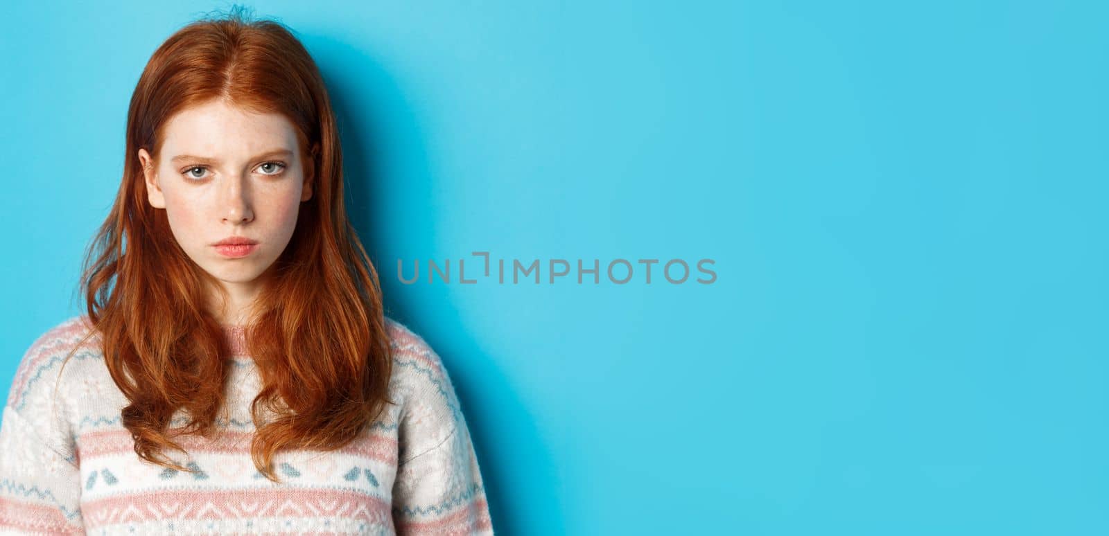 Close-up of angry redhead teenage girl looking pissed-off at camera, frowning and sulking mad, standing over blue background by Benzoix