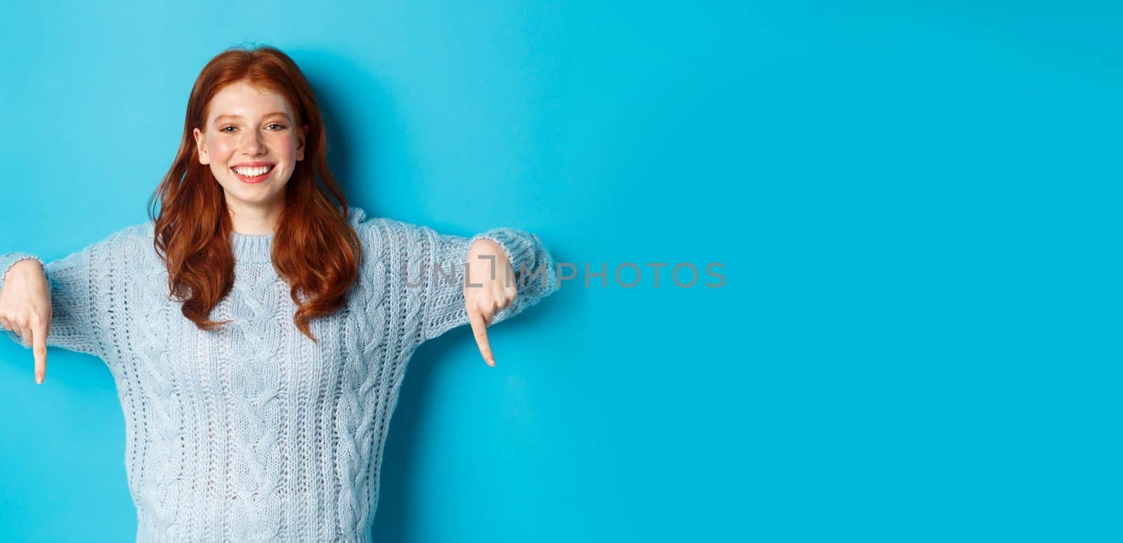 Winter holidays and people concept. Cheerful redhead woman in sweater pointing fingers down, smiling pleased at camera, showing promo, blue background by Benzoix