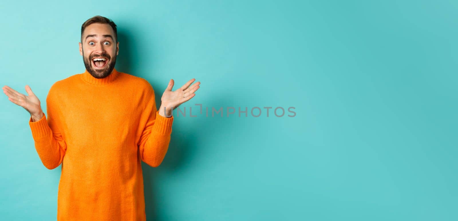 Surprised man raising hands up, looking at something amazing, standing over turquoise background in winter sweater.