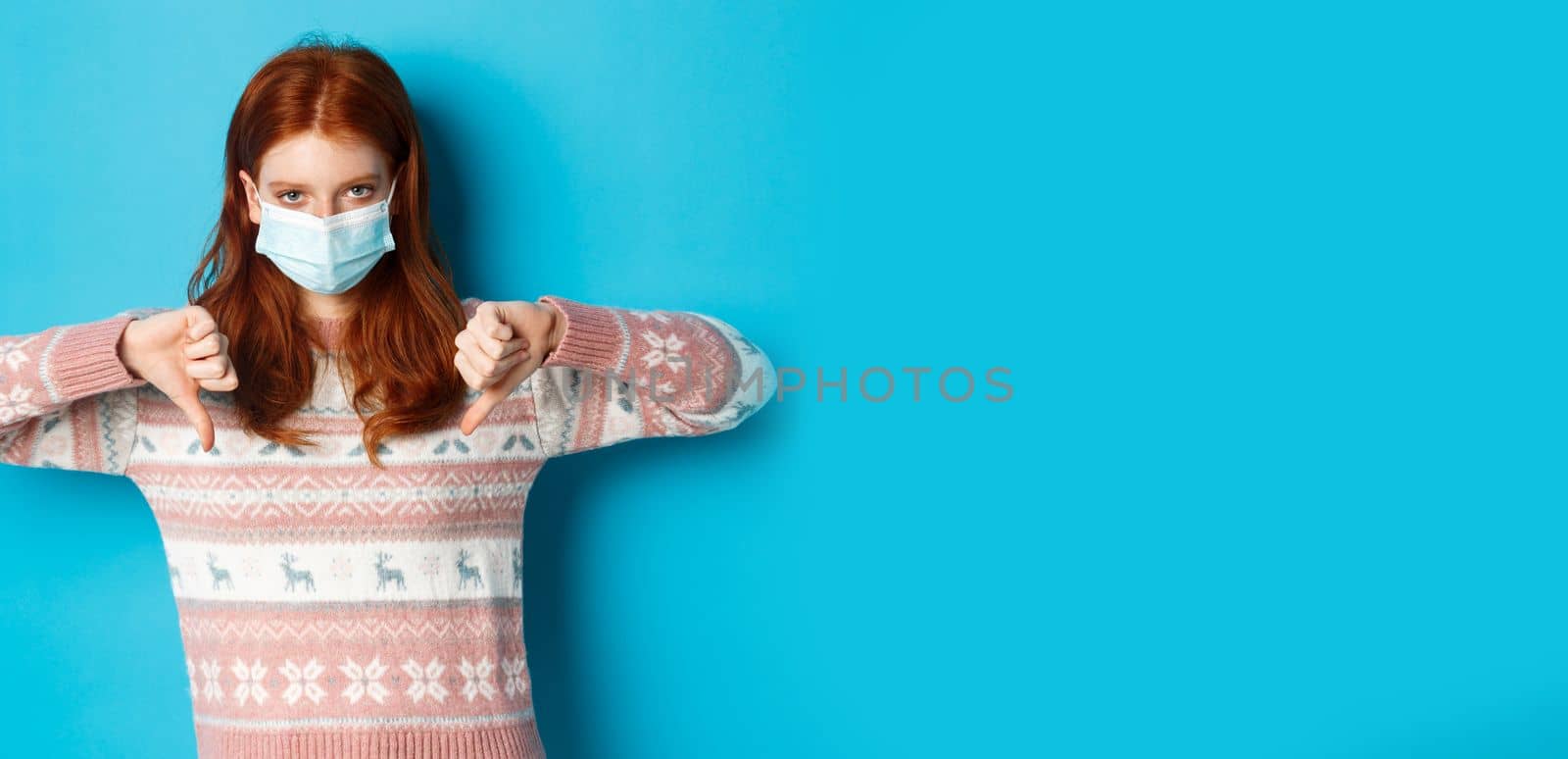 Winter, covid-19 and pandemic concept. Upset and angry redhead girl in face mask showing disapproval, thumbs down in dislike, standing over blue background.