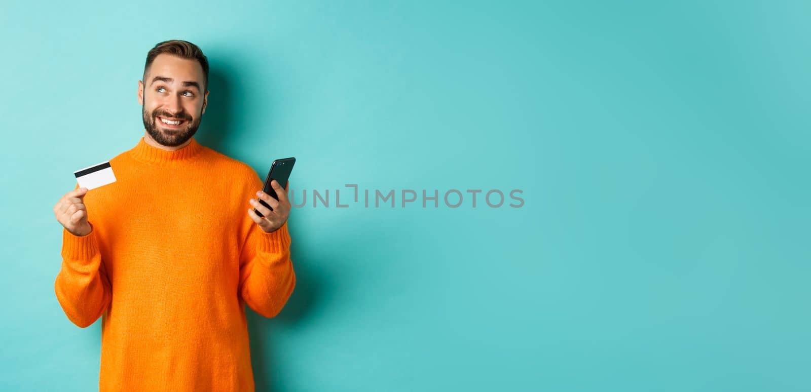 Online shopping. Handsome man thinking, holding smartphone with credit card, paying in internet store, standing over light blue background.