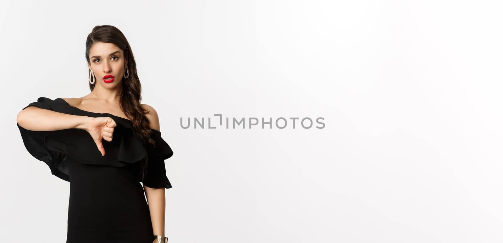 Fashion and beauty. Disappointed sassy woman in black dress, showing thumbs down, dislike something bad, judging over white background by Benzoix